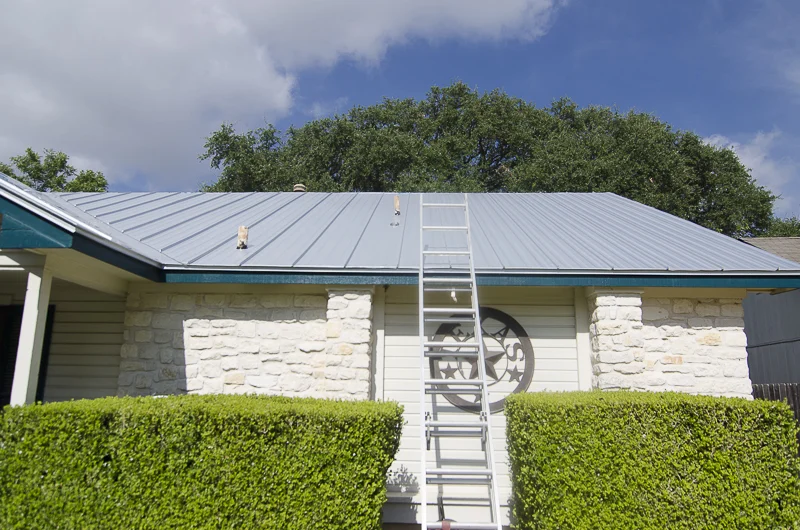 A new metal roof on a tan stone house with a ladder against it and bushes in front.